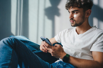 Young serious man wearing casual clothes using mobile cell phone browsing the internet sitting on sofa indoors at home on weekends. People leisure concept. - obrazy, fototapety, plakaty