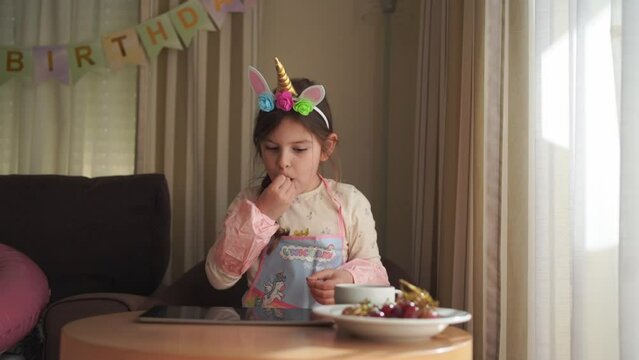 A young girl in a unicorn headband contemplates a unicorn picture on a birthday card at a themed party, with a bowl of grapes nearby.