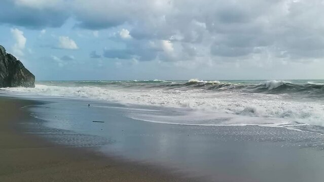 Spiaggia solitaria al rallentatore con il mare agitato: le onde si infrangono sulla riva e sulla roccia in lontananza