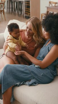 Vertical video of same sex female couple or friends playing with baby sitting on sofa at home together - shot in slow motion