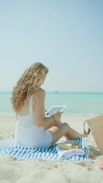 relaxed modern middle aged woman on the ocean shore with straw bag and striped towel using applications on tablet PC.