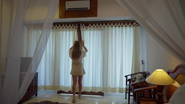 Woman going out balcony, enjoying view. Young woman opens curtains and looks to the beautiful landscape sea from villa balcony, enjoying rest. Vacation summer morning, resting, hotel relax concept.