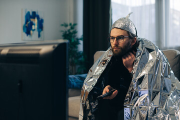 A young man wearing a tinfoil hat and wrapped in a tinfoil blanket is sitting on the couch in front...