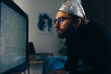 Conspiracy Theory on Screen: Man in Tin Foil Hat and Blanket Observing TV Interference - obrazy, fototapety, plakaty