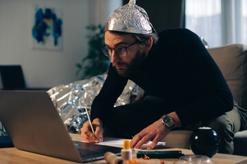 Conspiracy theory. A man in a tinfoil hat looks for signs while watching a video on a laptop. - obrazy, fototapety, plakaty