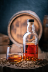 A glass of whiskey with bottle on wooden board. Oak barrel on background. Barley grains and barley ears on a foreground - 767290057