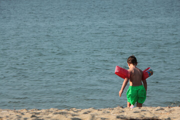 blonde boy walking towards the sea on the beach. boy in green shorts with red inflatable water armbands. Rear view of child with armbands walking on beach on summer holiday.