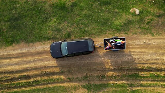 Aerial high angle top down shot of a black car towing a small trailer with two off road cross morotbikes along a windy dirt road. Drone footage of vehicle with a trailer behind going to sports event.
