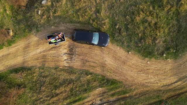 Aerial high angle top down shot of a black car towing a small trailer with two off road cross morotbikes along a windy dirt road. Drone footage of vehicle with a trailer behind going to sports event.
