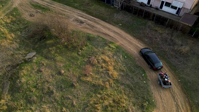Aerial high angle top down shot of a black car towing a small trailer with two off road cross morotbikes along a windy dirt road. Drone footage of vehicle with a trailer behind going to sports event.
