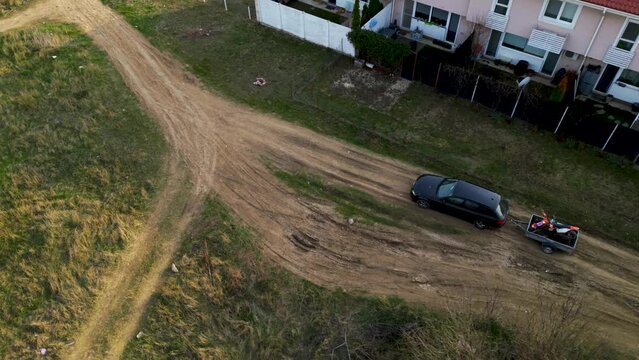 Aerial high angle top down shot of a black car towing a small trailer with two off road cross morotbikes along a windy dirt road. Drone footage of vehicle with a trailer behind going to sports event.
