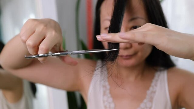 Young adult authentic asian woman self cutting bangs haircut with scissors. Background on day with nature light.
