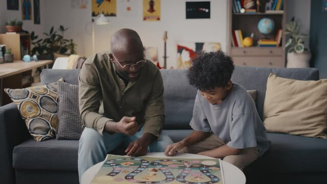 African American father and little son sitting together on couch in living room and talking while playing board game together at home