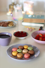 Plate of pastel macarons, cookies and chocolate, cup of tea of coffee, glass of bubble water, various berries, books and accessories on the table. Selective focus, pastel colors.