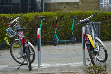 City bikes and electric scooters in the background parked in front of a shopping mall, early spring, eco-friendly transport in the city for short distances, car alternative in the city. - obrazy, fototapety, plakaty