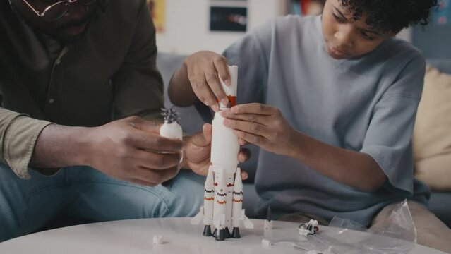 Little African American boy building rocket model with help of father while spending day together at home