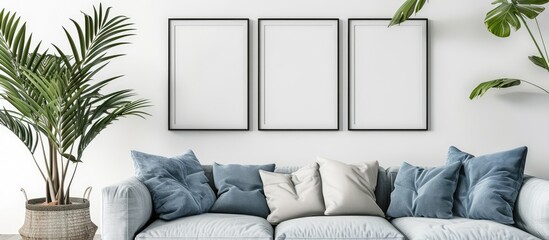 Blank frames on a white wall in a modern Scandinavian living room with a sofa, cushions, and a potted palm plant, featuring mockups of poster prints to showcase a home staging concept.
