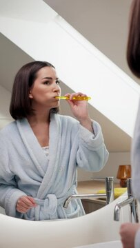 A woman in a bathrobe brushing her teeth in front of a bathroom mirror