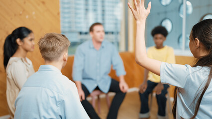 Smart girl raising hand to ask question during doing group therapy. High school student put hand up...