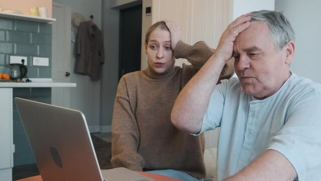 Daughter and elderly father shocked looking at laptop and touching their heads, failure with computer in kitchen at home. Frustrated old man and young woman amazed with problems