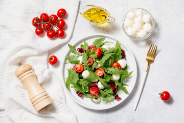 Fresh green salad made with mozzarella cheese, cherry tomatoes, arugula and olive oil on white stone table background. Healthy food and diet concept. Flat lay, top view, copy space.