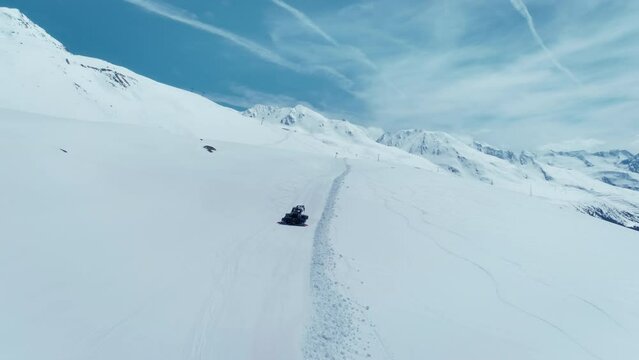 Drone follows snowcat plowing snow on ski slopes of ski resort on sunny day. Beautiful mountain landscape in Austrian Alps. Snowcat groomer prepares piste for skiing season. Winter resort preparation