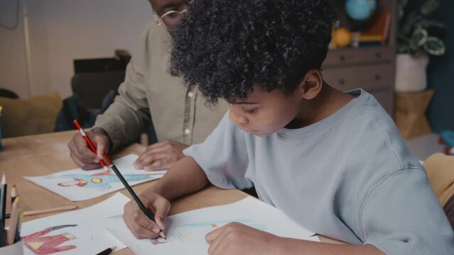 Little African American boy with curly hair sitting next to dad and drawing with colored pencil on paper during leisure time at home