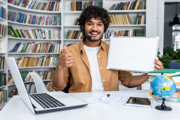 Smiling indian man with wireless headset holding blank whiteboard and giving thumbs up in front of modern laptop with various items on desk. Background of bookshelves and home office.