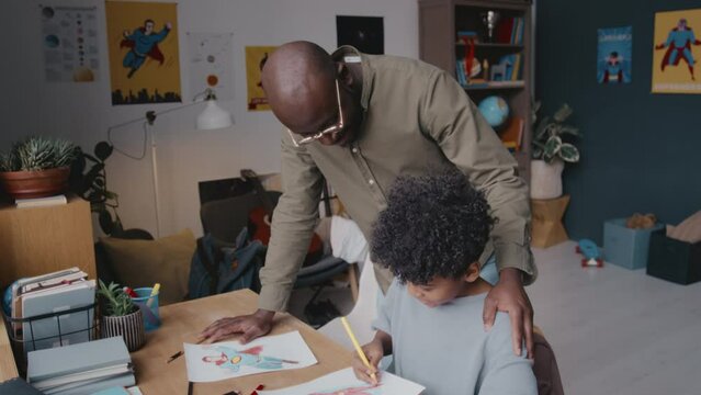 Black father walking to little kid drawing at desk in living room, looking at picture and praising him