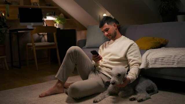 Young man holding mobile phone while sitting on carpet with dog and caresses his pet