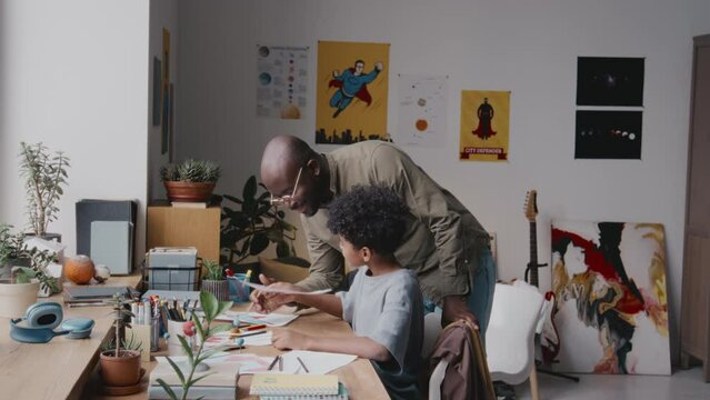 Little African American boy drawing at desk in kids room while dad walking to him, watching picture, praising son and ruffling his hair