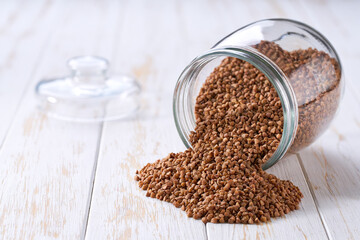 Raw buckwheat spill out of a glass storage jar on a white wooden table.