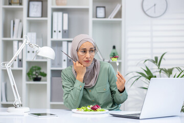 Front view of upset female sitting at desk with modern laptop and holding fork for eating salad. Frustrated arab woman feeling overeating and looking at dish with disgust in office environment.