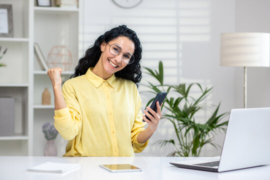 Portrait of beautiful woman in glasses sitting at personal workplace with mobile phone and expressing positive emotions. Smiling caucasian female doing winner gesture and looking at camera.
