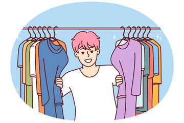 Young Cheerful man in t-shirt smiling peeking out from behind hanger for collection of clothes choosing new look. Guy with pink hair is standing in clothing store wanting to buy fashionable outfit