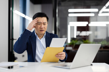 Shocked chinese male holding envelope with letter inside and reading important paper message at personal workplace. Man receiving surprise news while working at portable laptop at office.