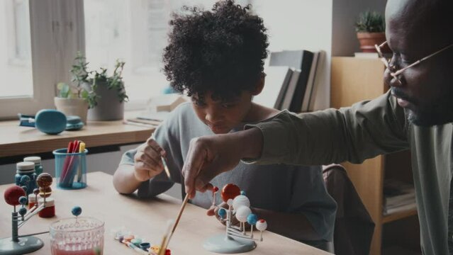 Little African American boy painting planets on DIY solar system model with assistance of father while doing craft project for elementary school at home