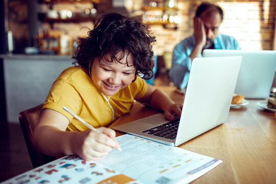 Happy child doing homework on laptop while father works in background at home
