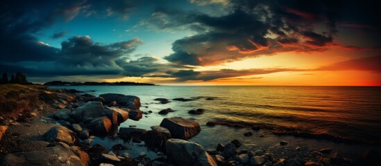 A picturesque sunset casting a golden glow over a tranquil lake, with rocks scattered along the shore in the foreground, under a colorful sky - obrazy, fototapety, plakaty