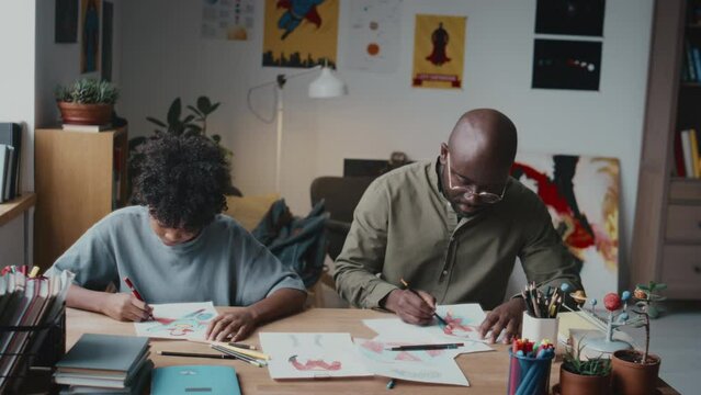 African American dad and little son sitting at desk in living room, using colored pencils and talking while drawing together during leisure time at home