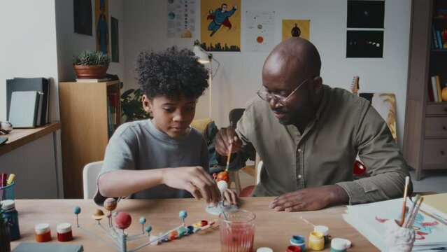 Little Black boy and his father sitting at desk in living room and painting planets on DIY solar system model while doing crafts together at home