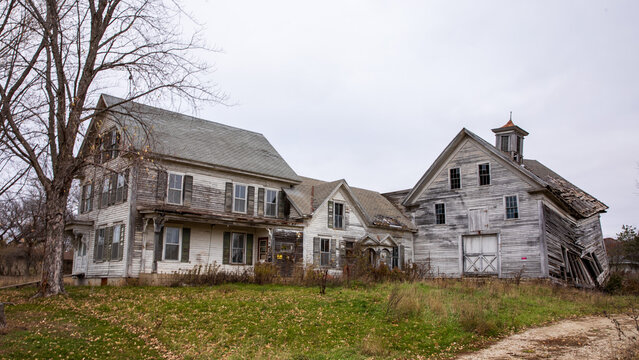 Abandoned home in rural setting. Country estate in ruin.  Pale sky over dilapidated buildings in countryside with open tan grassy field. 