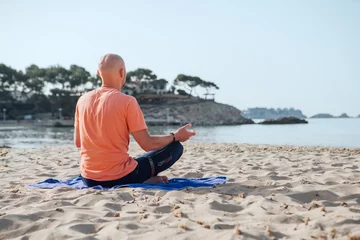 Poster Im Rahmen Calm man sitting in lotus position, doing deep breathing exercises and meditating in early morning hours on the sandy beach with calm sea waves. Mental health, people relaxing, traveling concept. © Soloviova Liudmyla
