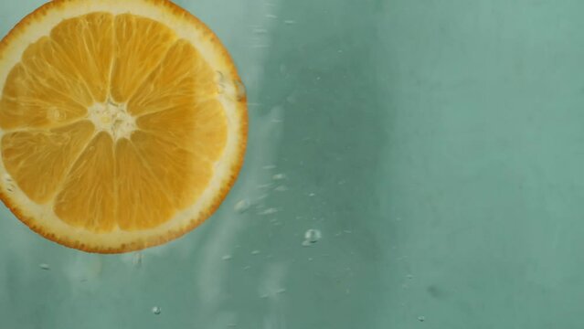 slice of orange drops into water causing playful splashes. As orange slice hits water it creates ripples that dance across surface as it sinks deeper into water leaving behind trail of citrus essence.