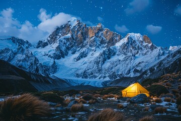 Illuminated yellow tent stands out against a backdrop of snow-covered mountains under a starlit sky