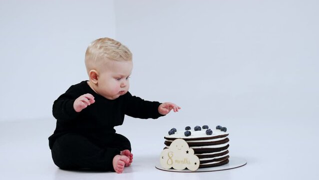 Adorable blond baby with cute plump cheeks sits at the big cake. Child wants to touch the dessert but changes his mind. White backdrop.