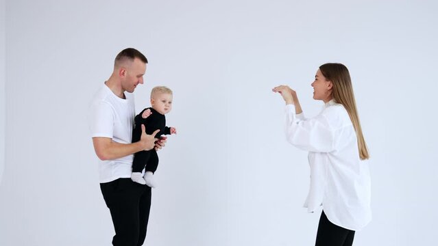 Caucasian man in white t-shirt holding baby boy in black suit. Mother amuses baby and tickles him but the kid stays serious. White backdrop.