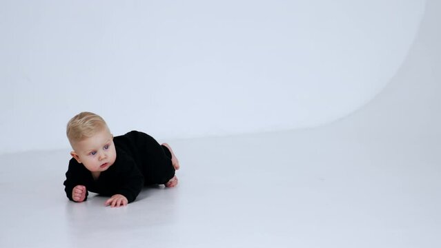 Little infant baby boy in black suit crawling by the floor. Adorable blond child at white backdrop.