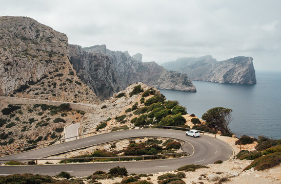Fototapeta Lonely small white car driving by serpentine curved asphalt mountain road near Lighthouse of Cap de Formentor with beautiful seascape with rocky coast. Mallorca Island, Balearic Islands, Spain.