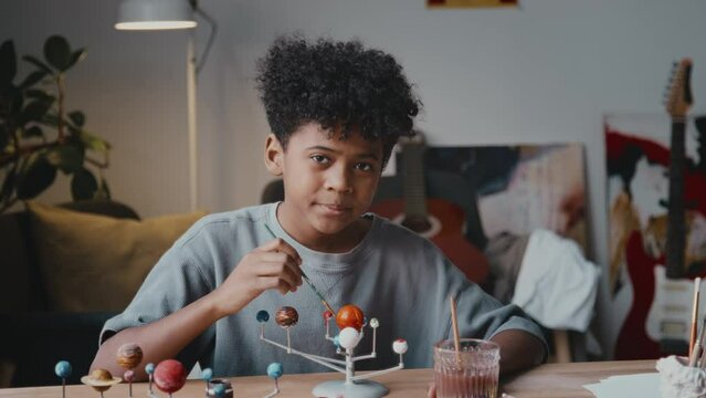 Portrait of little African American boy painting DIY solar system model and smiling on camera while doing craft project for school at home